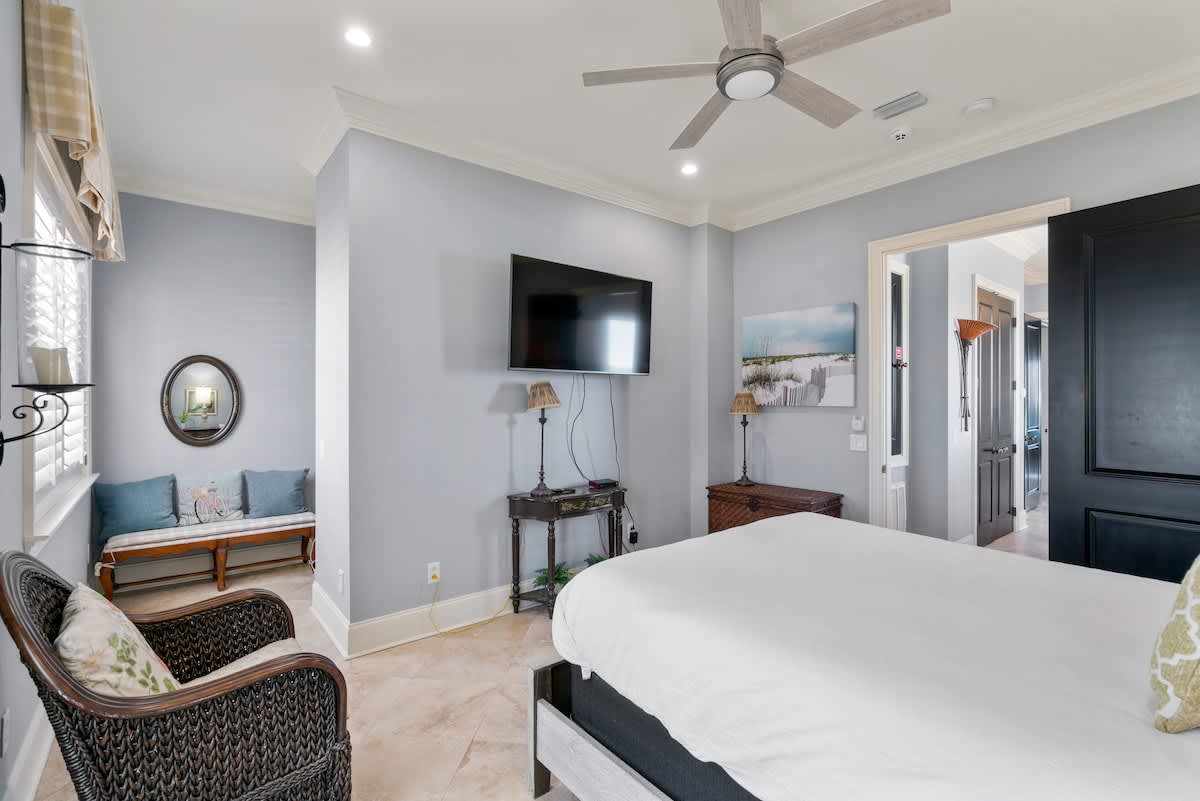 Bedroom with light blue walls and travertine tile flooring, featuring a queen bed and a wall-mounted TV. An alcove with a wooden bench and blue pillows is visible, along with a wicker chair and decorative wall sconce.