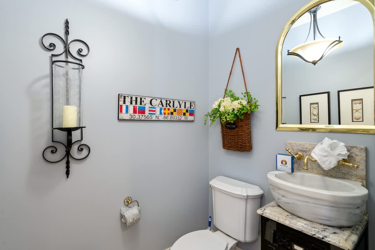 A powder room with light blue walls, featuring a unique marble vessel sink on a dark vanity and an arched gold-framed mirror. A decorative wall sconce and a nautical-themed sign add character.