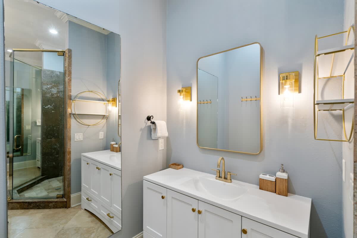 A vanity area in a light blue bathroom with travertine flooring, featuring a white cabinet, gold faucet, rectangular gold-framed mirror, and gold wall sconces. A full-length mirror reflects the shower.