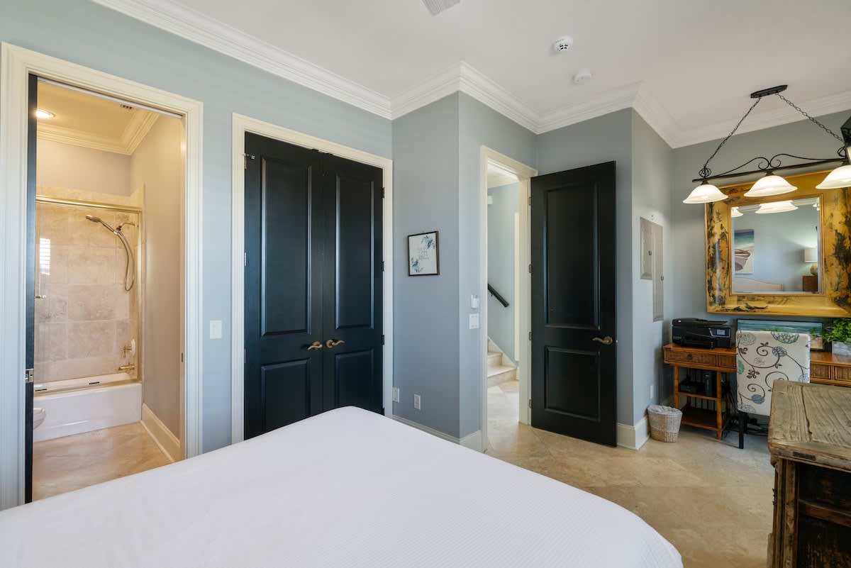 This bedroom features light blue walls, tile flooring, and black double doors, with an open doorway leading to an en-suite bathroom. A staircase is visible through another open door, suggesting a multi-level property.