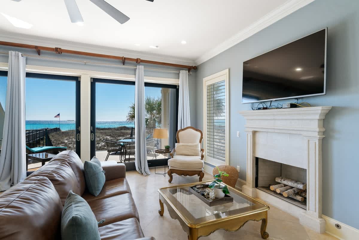A cozy living room featuring a brown leather sofa, a gold-framed coffee table, a stone fireplace, and a wall-mounted TV, with direct access to an outdoor patio offering beach and ocean views.