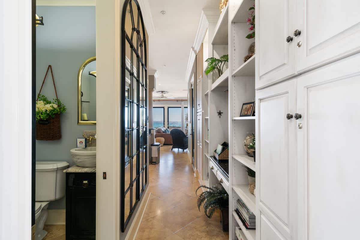 A bright hallway with light tile floors and white built-in cabinetry, leading towards a living area with ocean views. A glimpse of a bathroom with a unique vessel sink is visible to the left.