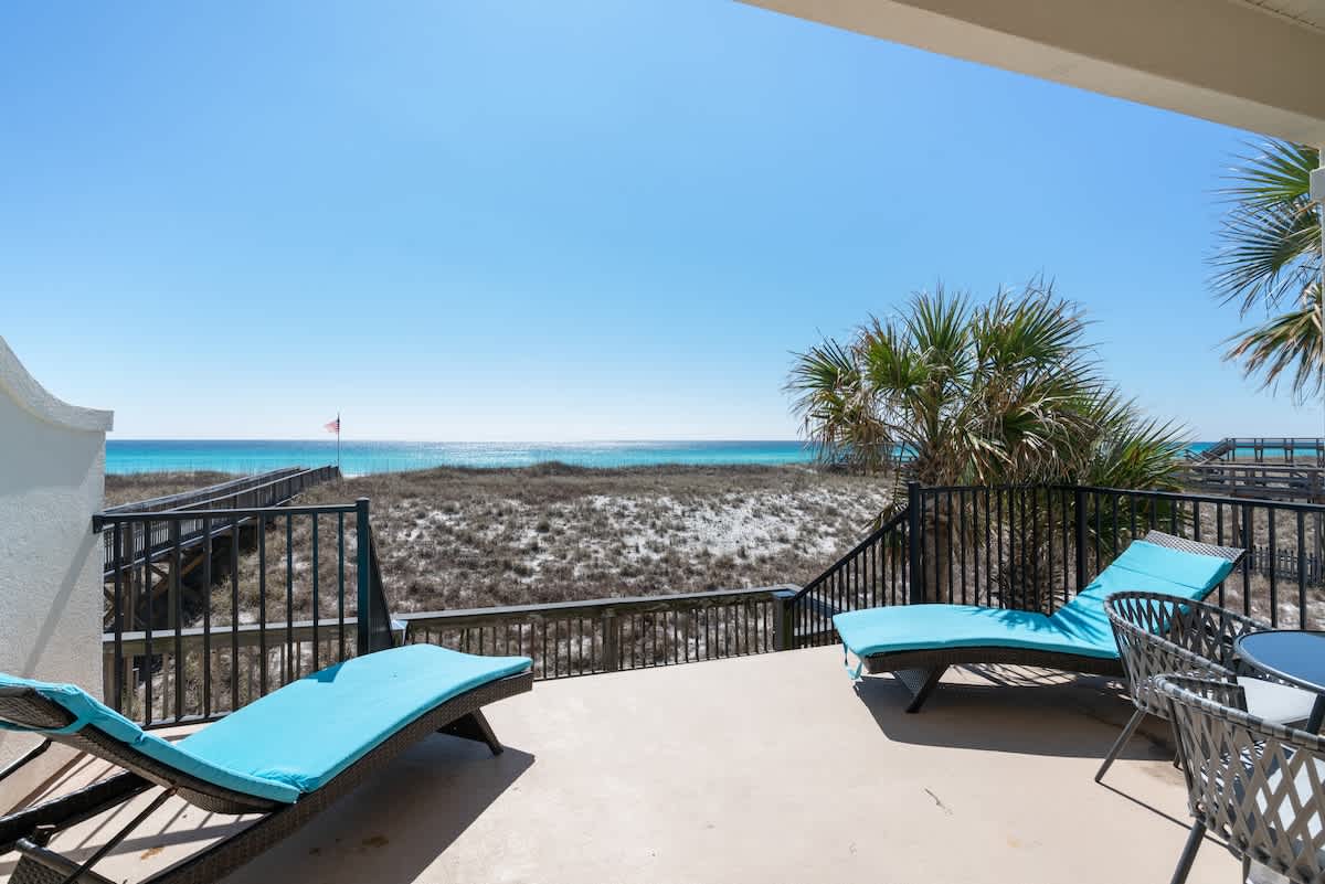 A spacious outdoor balcony featuring two blue chaise lounge chairs and a small round table with chairs, offering a stunning view of the white sand beach, dunes, and clear blue ocean.