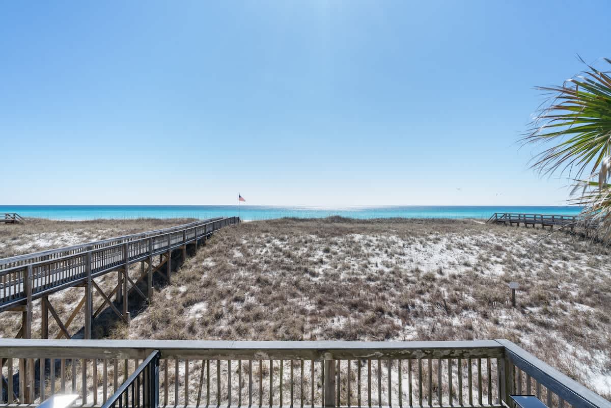 A wide view from a balcony showing a long wooden boardwalk winding through sand dunes towards the vibrant turquoise ocean, with a clear blue sky overhead.