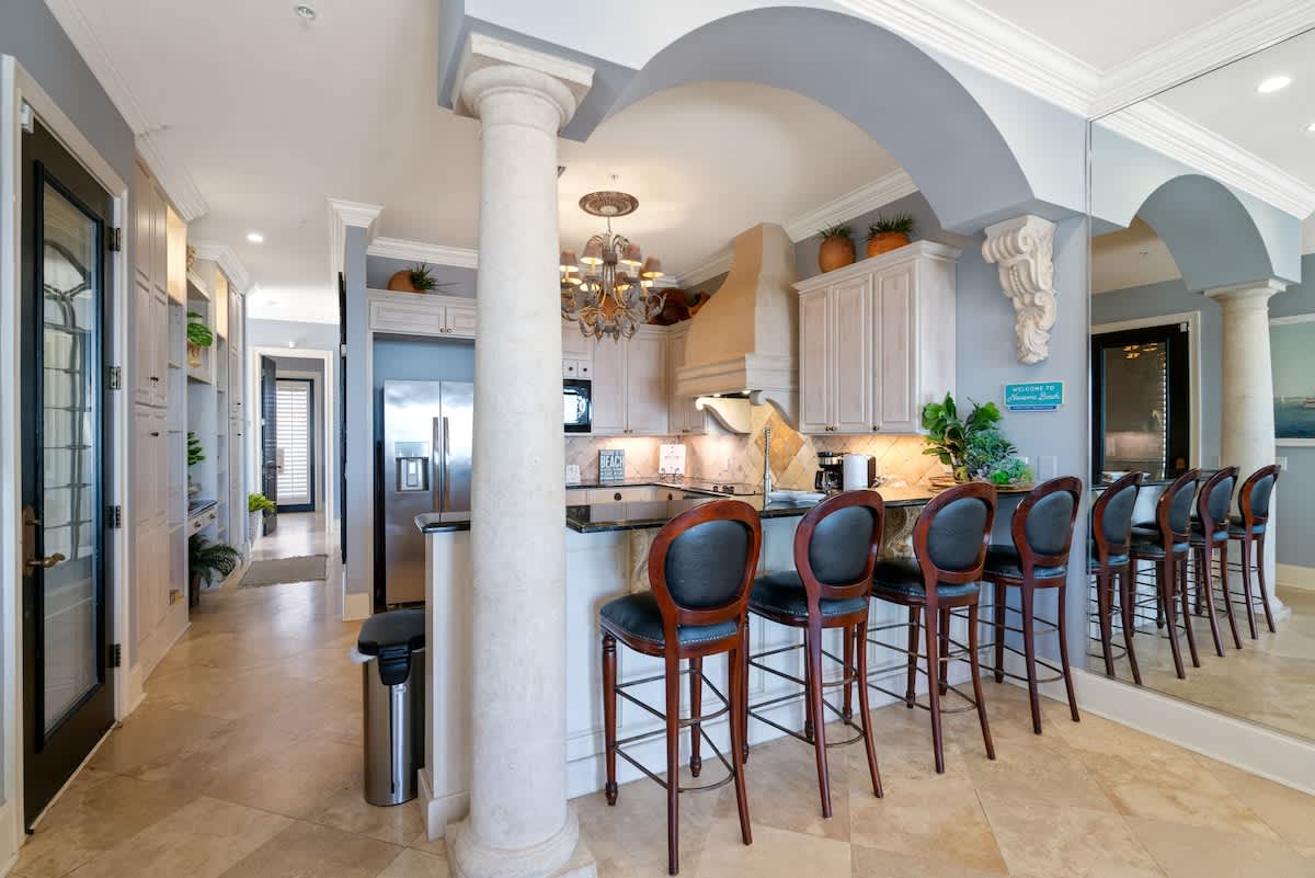 A spacious kitchen featuring light-colored cabinets, dark granite countertops, stainless steel appliances, and a long breakfast bar with multiple dark leather bar stools, framed by an elegant archway and column.