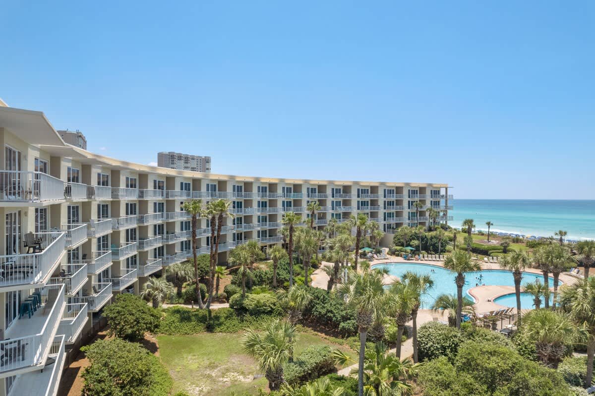 Sweeping aerial view of a curved resort building with many balconies, overlooking a large outdoor pool area with palm trees, and the vast expanse of the ocean and white sand beach beyond.