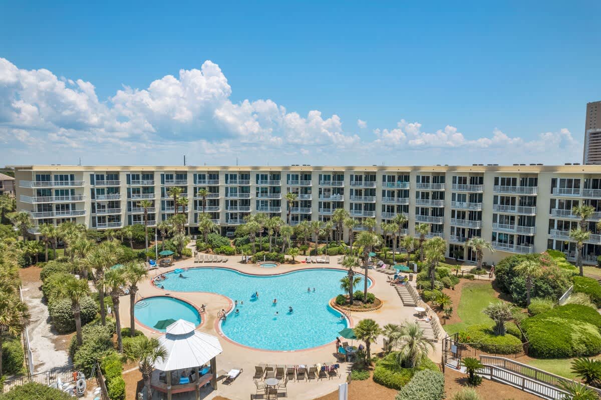 Wide aerial shot showcasing the expansive, freeform swimming pool with multiple sections and a hot tub, surrounded by lounge chairs, palm trees, and lush greenery, all set in front of a long, multi-story condominium building.