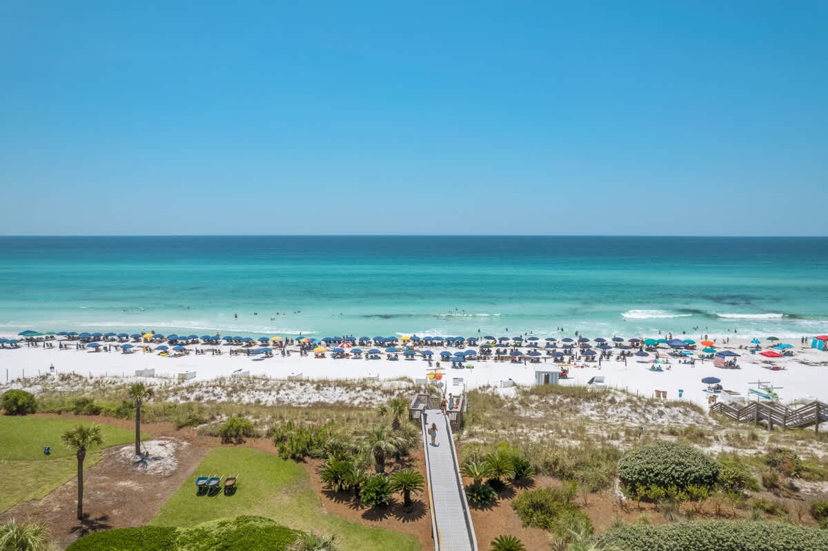 Elevated view of a pristine white sand beach dotted with numerous colorful umbrellas and lounge chairs, leading to the clear turquoise waters of the ocean under a bright blue sky, with a wooden boardwalk providing access from the property.