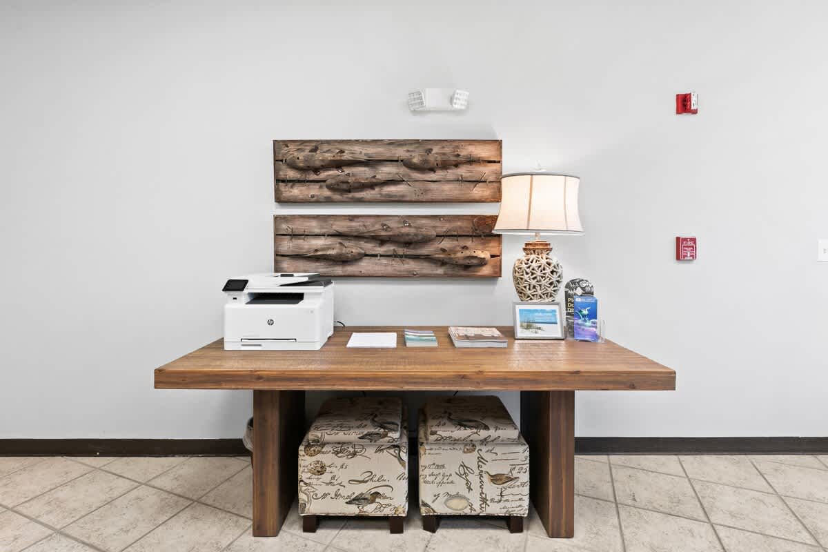 A dedicated business corner within the common area, featuring a rustic wooden console table with a printer, a lamp, and decorative wooden wall art. Two upholstered ottomans are tucked beneath the table.