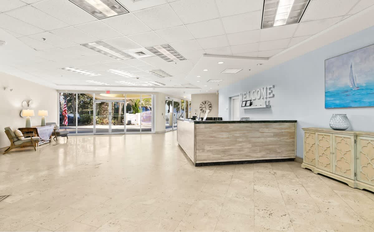 A wide view of the bright and spacious foyer, featuring a light wood reception desk with a dark countertop and a "WELCOME" sign. A comfortable seating area with modern chairs and a wooden console table is visible, alongside large windows offering natural light.