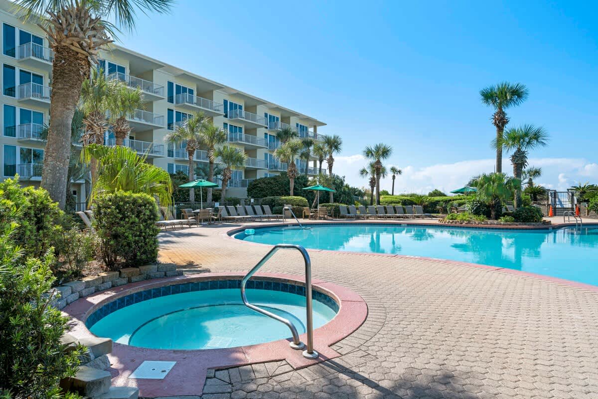 A large outdoor swimming pool with a separate, smaller circular hot tub, both surrounded by a textured paver deck and lush tropical landscaping with palm trees.