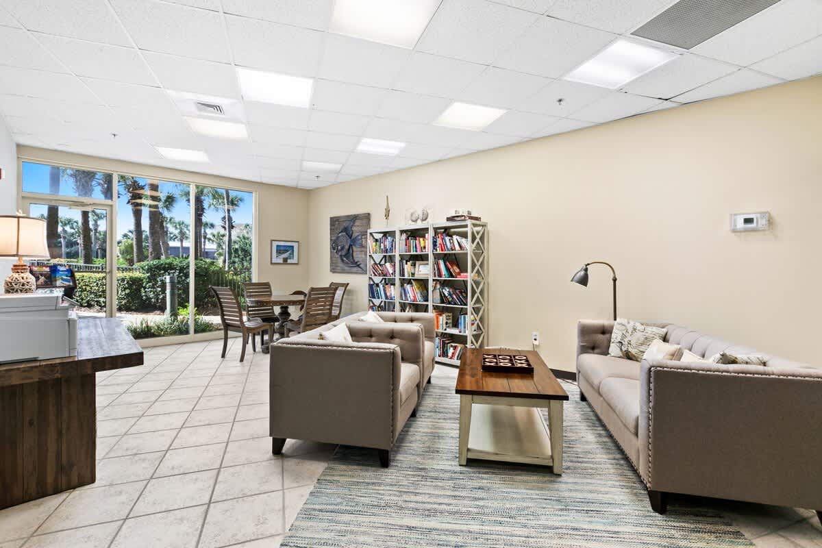 A spacious lounge area with light-colored tile flooring, featuring two beige sofas, a wooden coffee table with a Tic-Tac-Toe game, and a bookshelf filled with books. Large windows offer views of palm trees and outdoor seating.