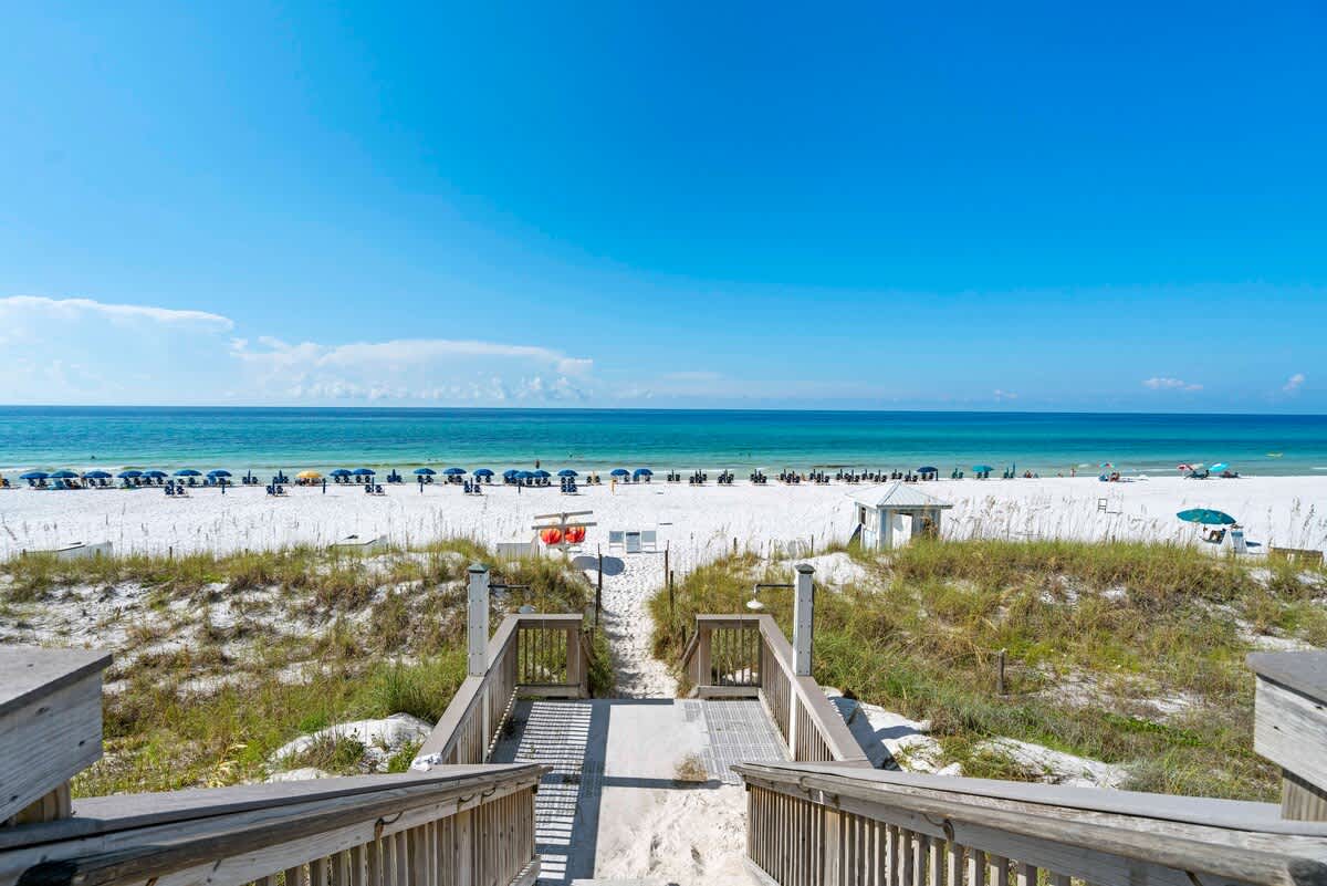 Wooden stairs lead down from a dune path to a wide, white sand beach dotted with blue umbrellas and lounge chairs, with the turquoise ocean stretching to the horizon under a clear sky.
