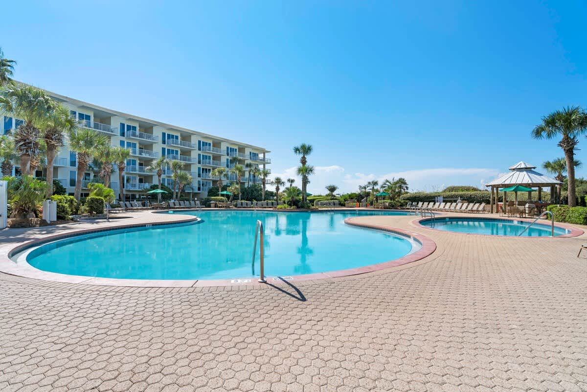 Wide view of a large, irregularly shaped outdoor swimming pool with clear blue water, bordered by a textured paver deck and surrounded by palm trees and resort buildings.