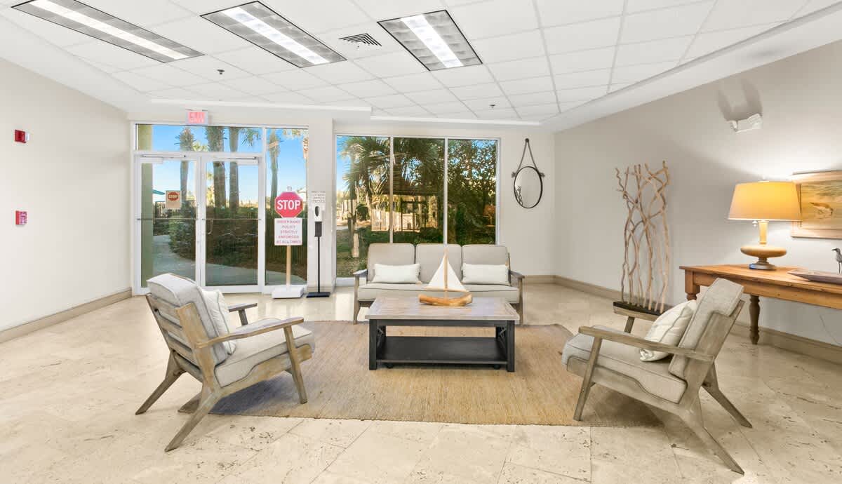 A comfortable seating arrangement in the foyer, featuring a light-colored sofa and two armchairs around a dark wood coffee table on a natural fiber rug. Large windows reveal lush palm trees outside, and a "STOP" sign is visible near the glass exit doors.