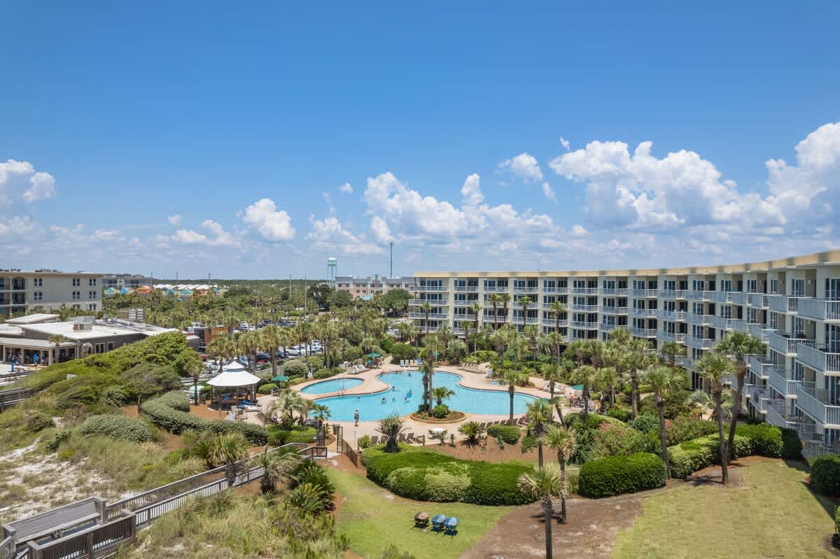 Elevated view of the condominium's large, freeform swimming pool area, surrounded by dense palm trees and manicured landscaping, with the multi-story building in the background and a water tower visible on the distant horizon.
