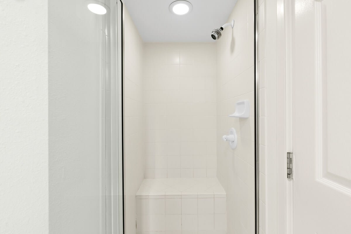 Clean, white-tiled walk-in shower with a built-in bench and a recessed light, featuring a simple shower head and a glass enclosure.