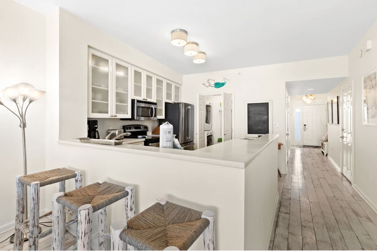Functional kitchen featuring a breakfast bar with woven bar stools, white cabinetry, and stainless steel appliances, with a hallway leading to the entrance and a laundry closet visible.