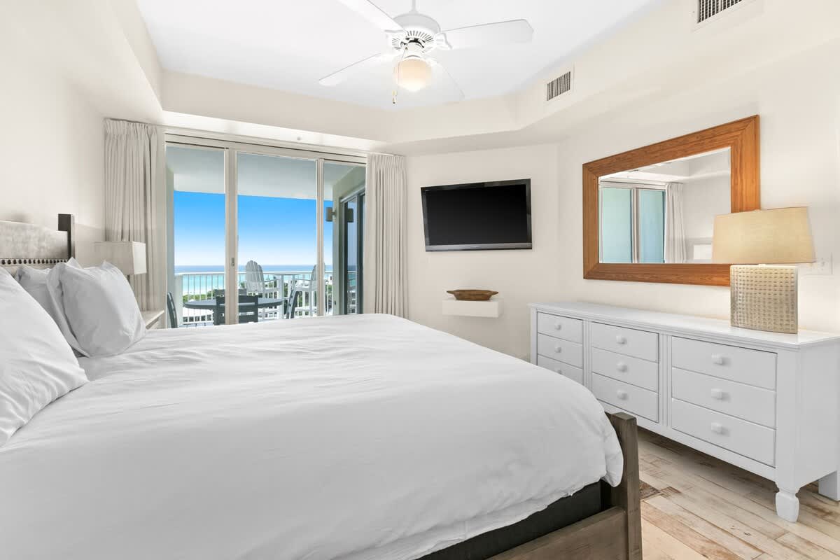 View of the master bedroom from the bed, showing a wall-mounted TV, a white dresser with a large wooden mirror, and sliding glass doors revealing a stunning ocean view from the balcony.
