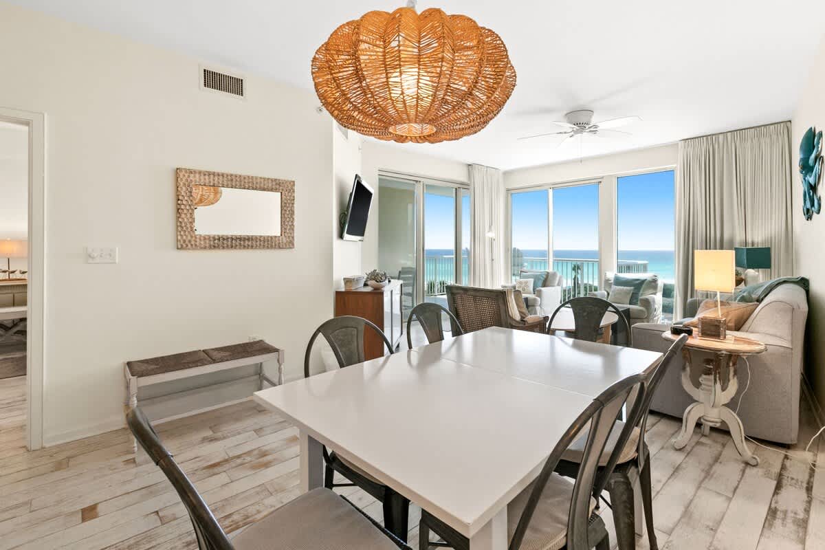 Bright dining space with a white table and metal chairs, complemented by a rustic wooden mirror and a bench against the wall, with the living room and ocean visible in the background.
