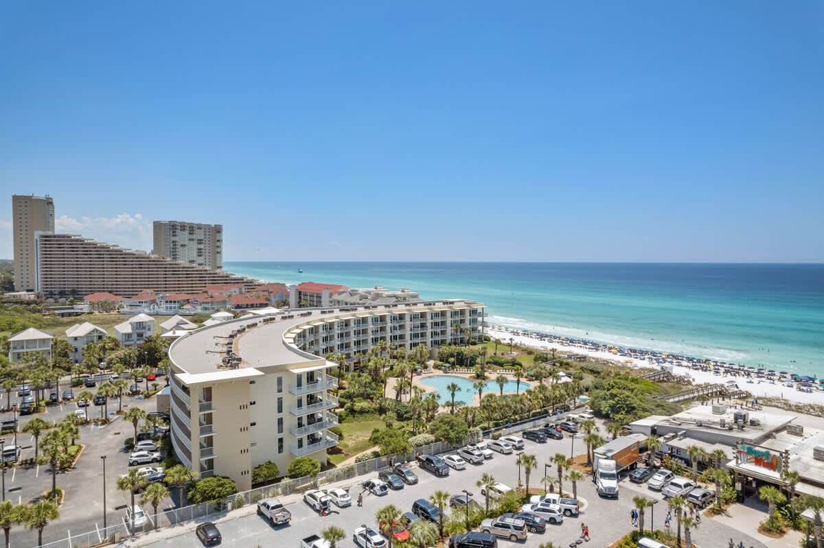 Broad aerial view capturing the curved condominium building, its large swimming pool area, and extensive parking facilities, with the white sand beach and turquoise ocean in the background, alongside other high-rise resort buildings in the distance.