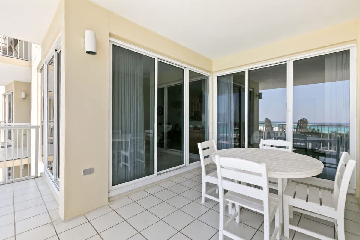 A large balcony with white tiled flooring, furnished with a white round dining table and chairs, offering ocean views and access to the interior through multiple sliding glass doors.