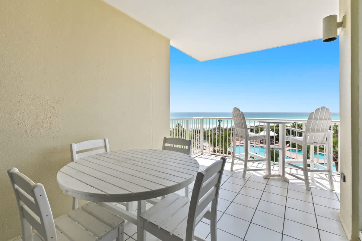 An expansive balcony overlooking the ocean and a resort pool, equipped with a white round dining table and chairs, plus a separate high-top table with two Adirondack-style chairs.