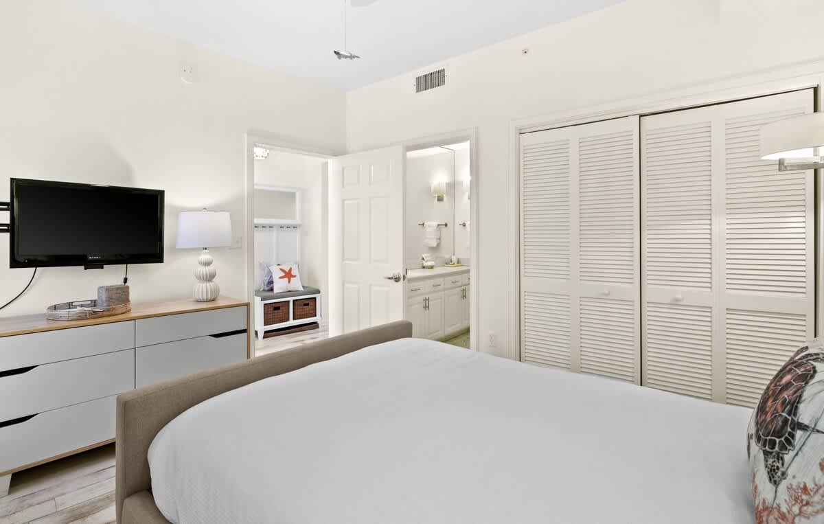 View of the second bedroom showing a dresser with a wall-mounted TV, a decorative bench with storage, and an open doorway leading to its en-suite bathroom.