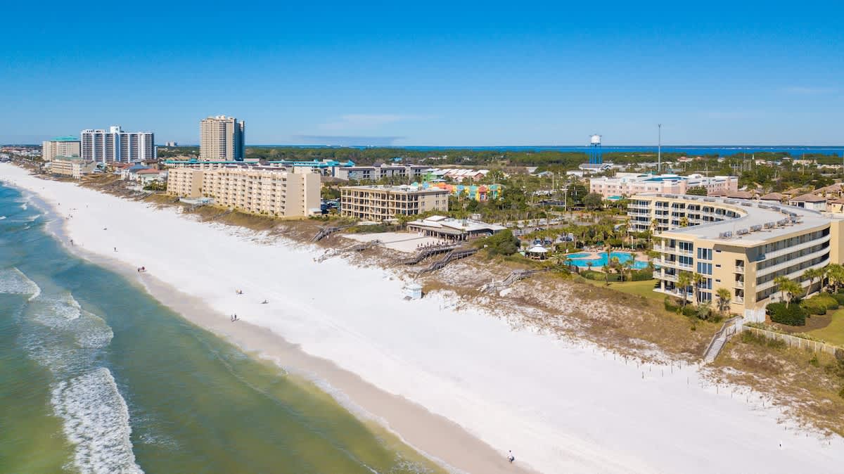 A wide aerial perspective captures the vibrant turquoise ocean gently lapping a long stretch of white sandy beach, bordered by numerous resort condominiums and lush coastal vegetation.