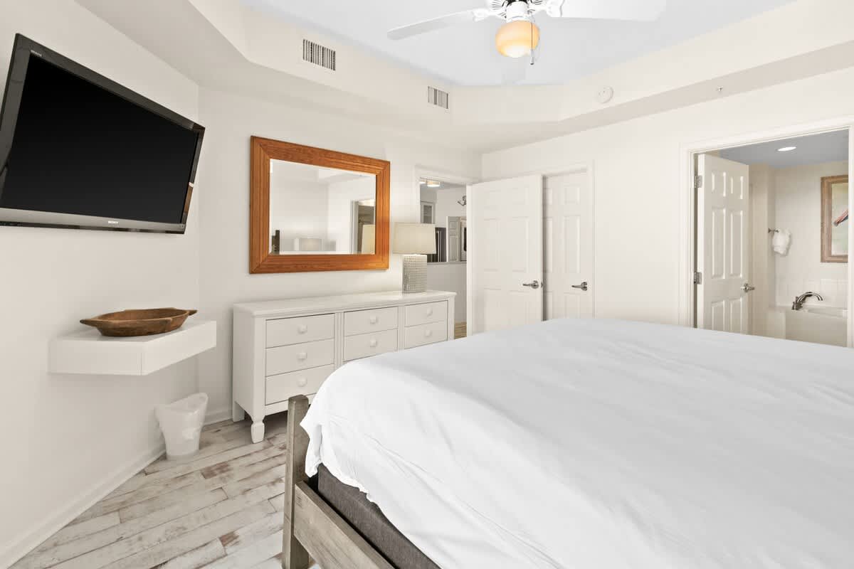 Corner view of the master bedroom with a large bed, a wall-mounted TV above a floating shelf, and an open doorway leading into the en-suite bathroom, featuring distressed wood floors.