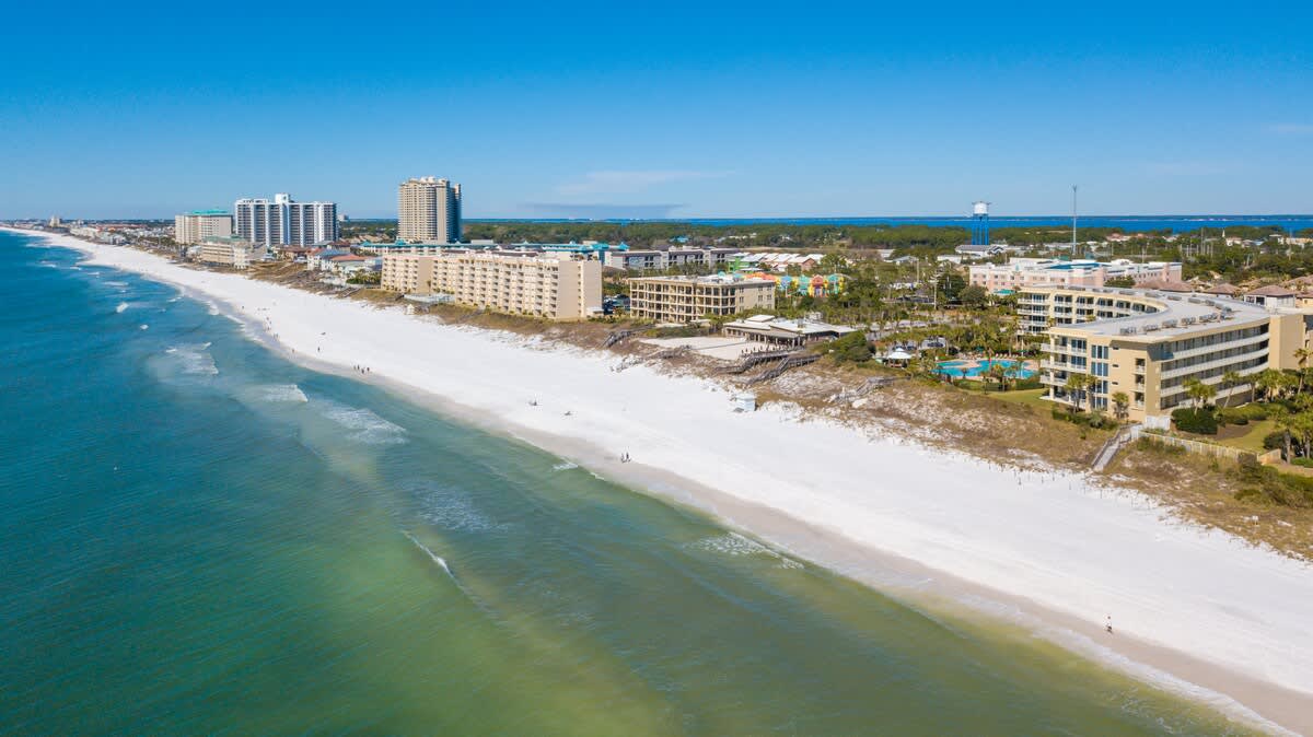 Expansive aerial panorama of a long, pristine white sand beach meeting the clear turquoise ocean, with several large resort complexes, including the featured condominium, stretching along the coastline under a bright blue sky.