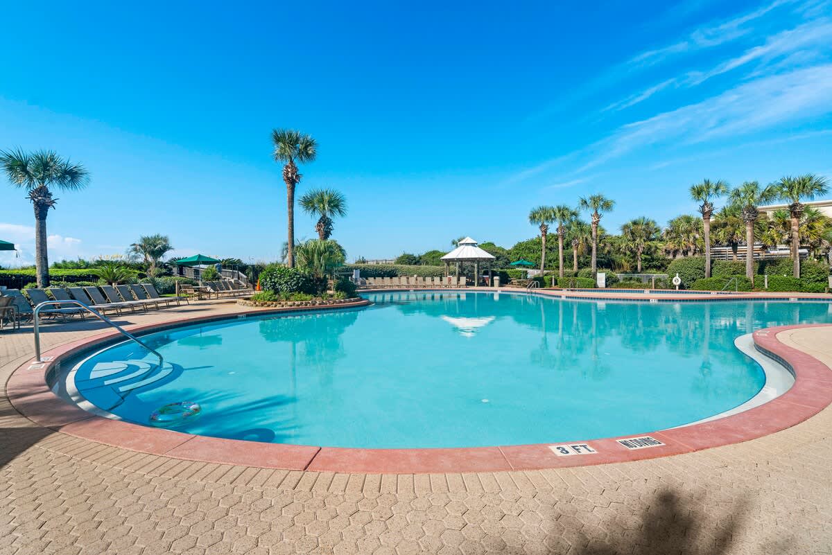 Expansive communal outdoor pool with clear blue water, surrounded by a textured paver deck, numerous lounge chairs, and lush palm trees under a bright blue sky.