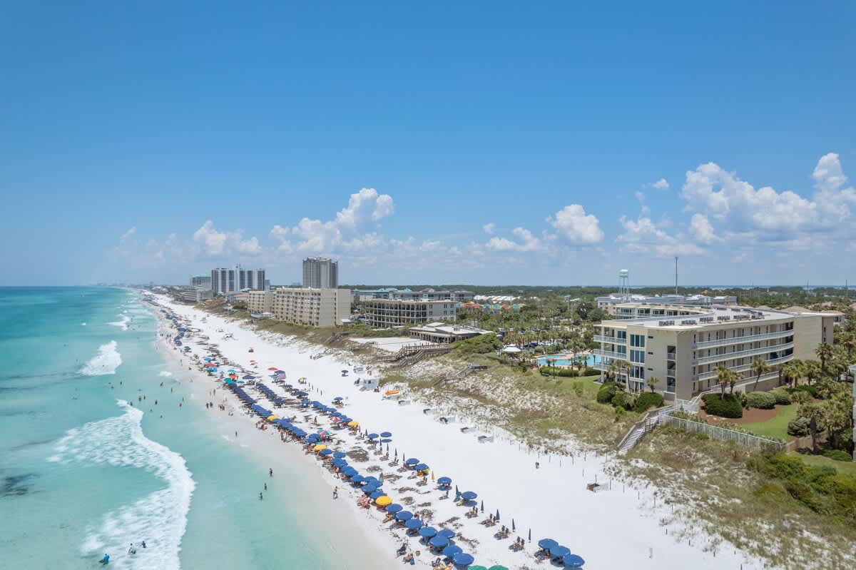 Panoramic aerial view of the vibrant coastline featuring a wide white sand beach, clear turquoise ocean, and a series of resort buildings, including the condominium, nestled among palm trees and lush dunes under a bright blue sky.