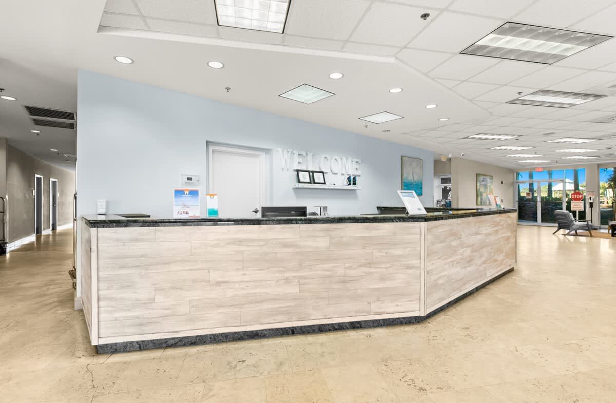 A modern reception desk with a light wood paneled front and dark countertop, featuring a "WELCOME" sign on the light blue wall behind it. The area is brightly lit by overhead fluorescent lights and has a light-colored tiled floor.