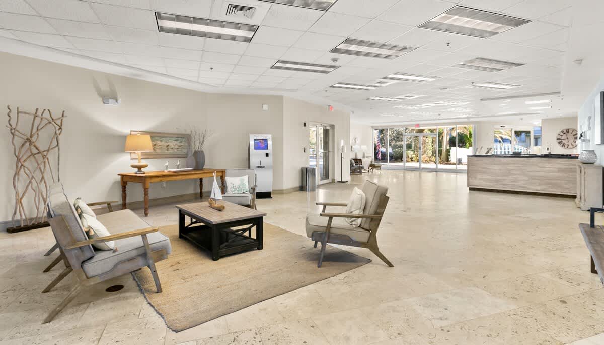 A panoramic view of the expansive foyer, showcasing multiple inviting seating areas with sofas and armchairs on rugs, and a wooden console table with lamps. The reception desk is visible in the background, along with a DVD rental machine and large windows.