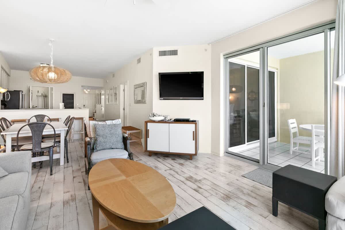 Open-concept living space with a wall-mounted TV above a wooden console, leading to a balcony with outdoor seating and a glimpse of the ocean beyond.