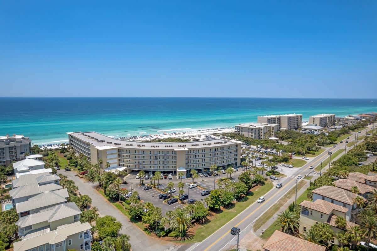 High aerial view showcasing the long, curved condominium building with its adjacent parking lot, a busy road, and residential homes, all situated near the expansive turquoise ocean and white sand beach under a clear blue sky.