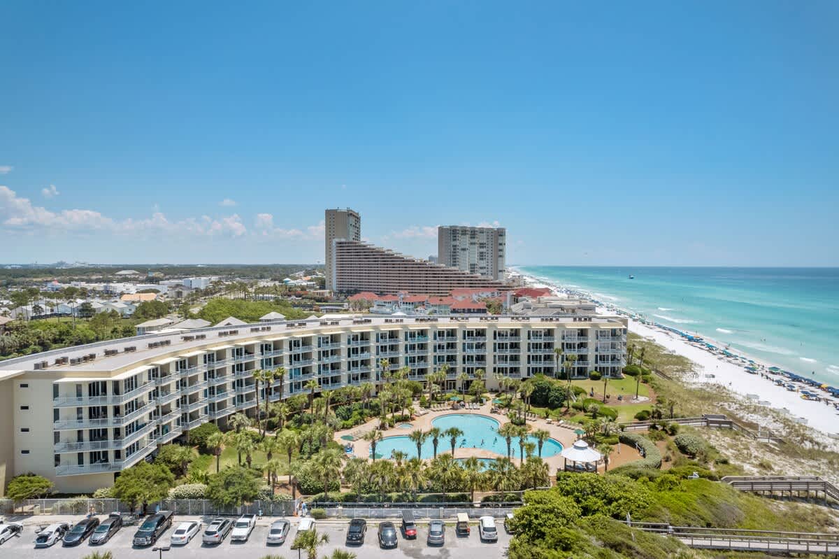 An aerial view showcasing a large resort building with multiple balconies, a sprawling communal pool area surrounded by palm trees, and a parking lot, all adjacent to a white sand beach and the ocean.