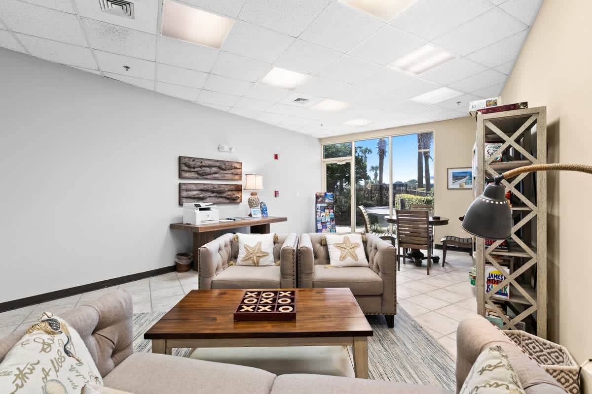 A comfortable lounge area with light tile flooring, featuring a sofa and two armchairs with decorative pillows, a wooden coffee table, and a bookshelf. A wooden console table with a printer and lamp is against a white wall, and large windows show outdoor views.
