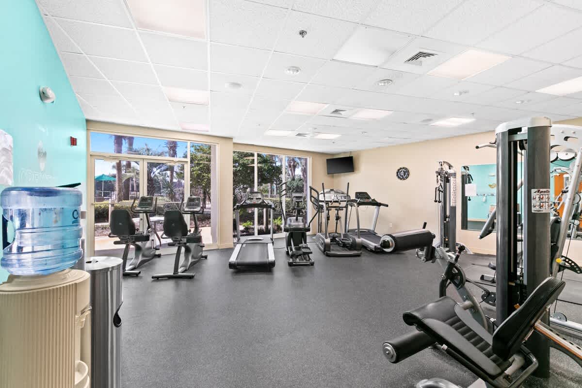A bright communal fitness center equipped with a variety of cardio machines including treadmills, ellipticals, and stationary bikes, alongside weight machines and large windows.
