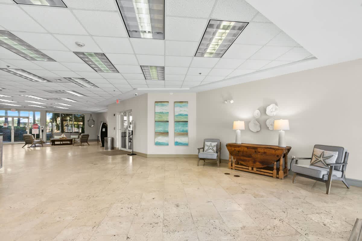 A spacious, brightly lit foyer with light beige tiled flooring, a drop ceiling with fluorescent lighting, and multiple seating areas featuring modern chairs and a wooden console table with lamps. Large windows at the far end offer a glimpse of the outdoors.