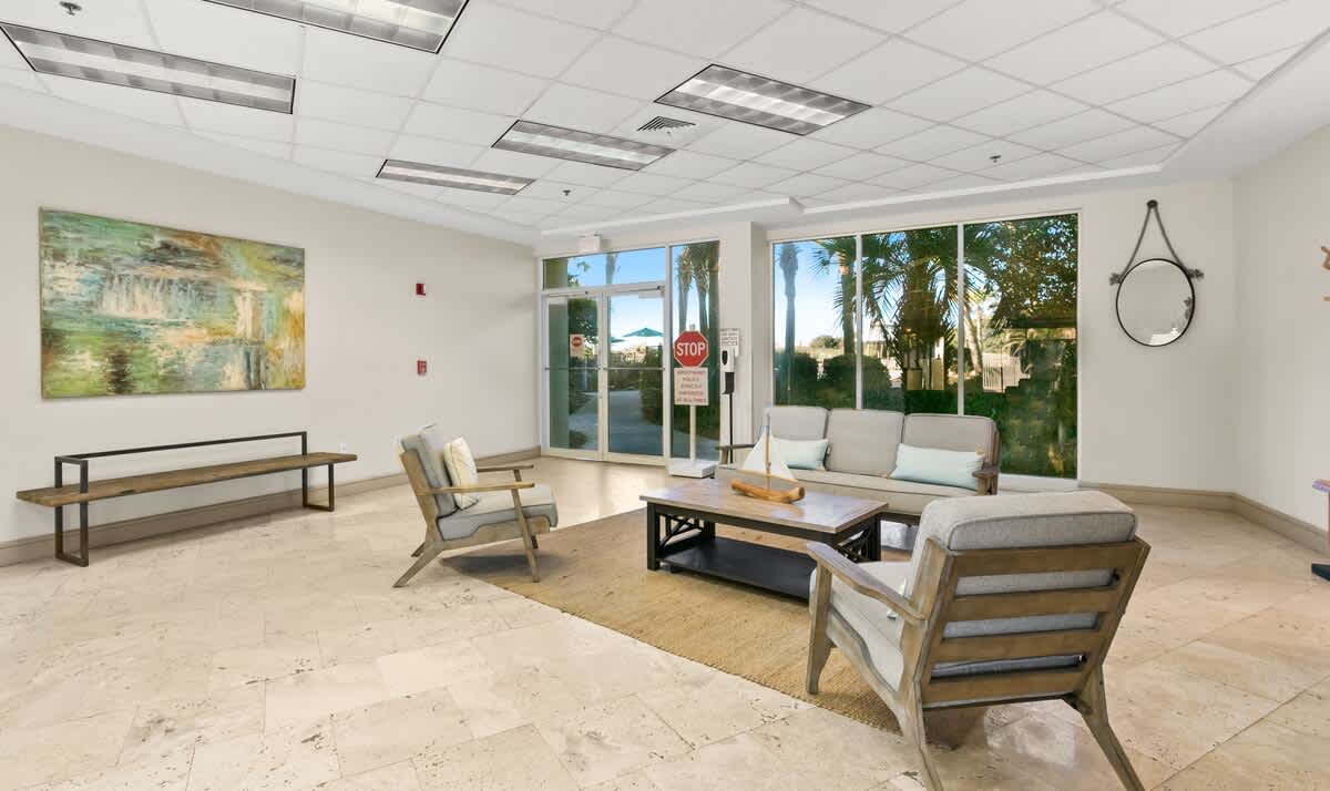 A welcoming seating area in the foyer with a light-colored sofa and two armchairs arranged around a coffee table on a textured rug. A large abstract artwork hangs on the wall, and a wooden bench is positioned beneath it, with large windows providing natural light.