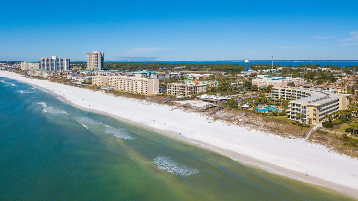 An expansive aerial view showcases a pristine white sand beach meeting turquoise ocean waters, with multiple high-rise and mid-rise resort buildings lining the coast under a clear blue sky.