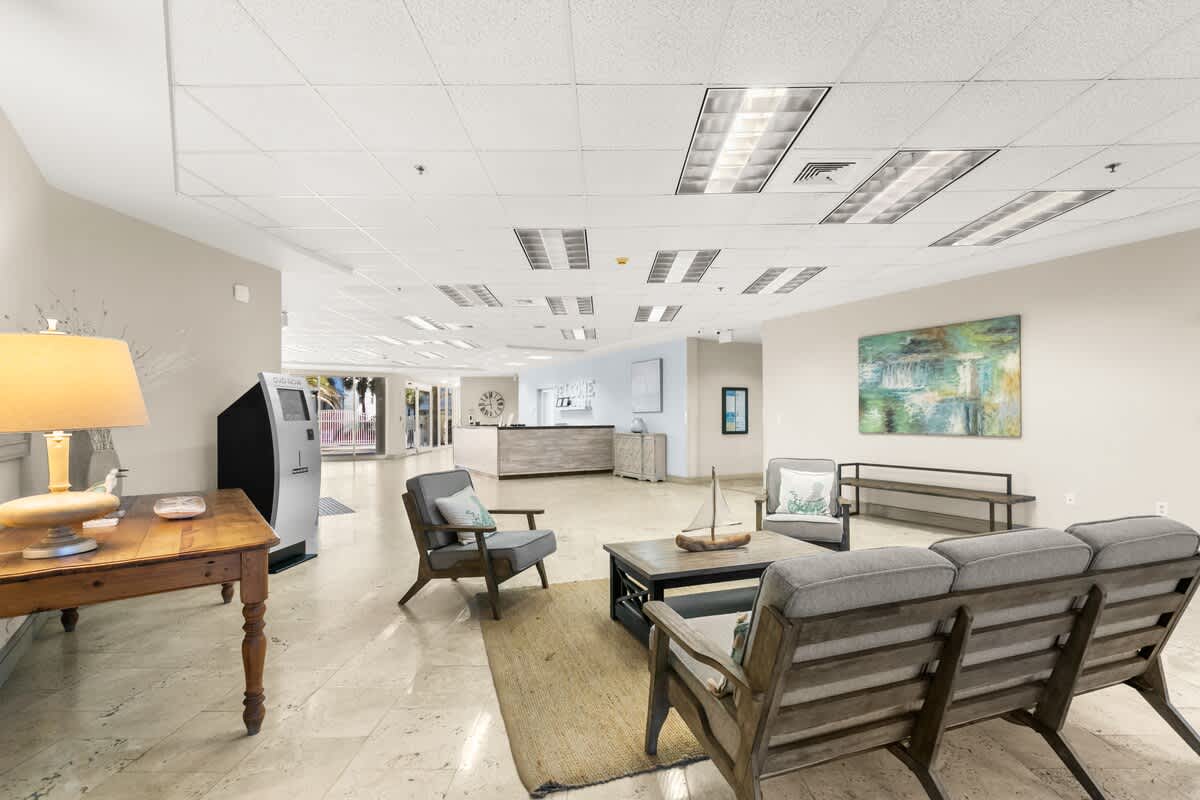 A comfortable seating area with a light-colored sofa and armchairs around a coffee table on a rug, facing towards the reception desk in the distance. An abstract artwork adorns the wall, and a DVD rental machine is visible to the left.