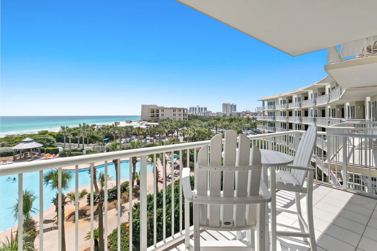 A wide-angle view from a balcony showcasing a large resort pool, numerous palm trees, and the expansive ocean, with white Adirondack-style high-top chairs and a small table.
