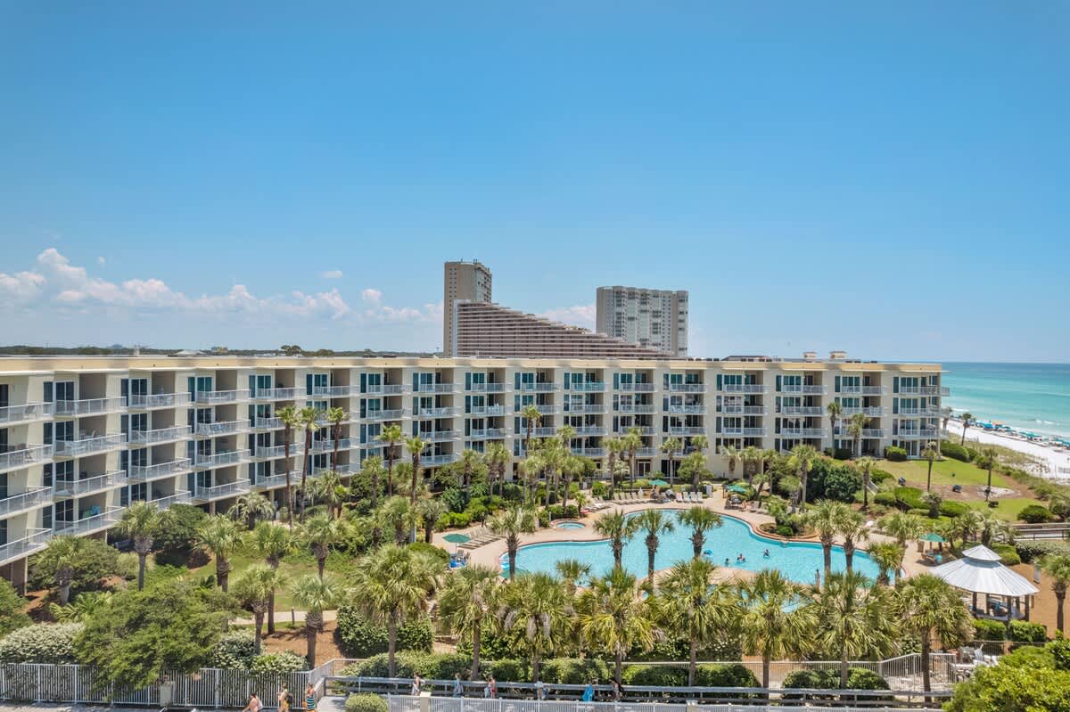 High-angle view of a large resort complex featuring a multi-story building with numerous balconies, a large outdoor pool, and abundant palm trees, situated near a white sand beach and the ocean.