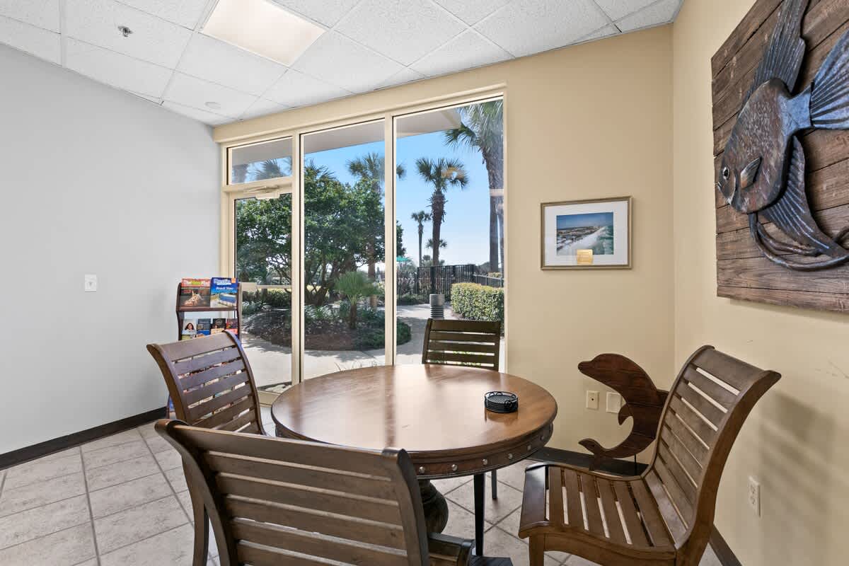 A small dining or game area with a round wooden table and four matching wooden chairs, situated next to large windows offering views of lush outdoor greenery. A large wooden fish decoration adorns the wall.