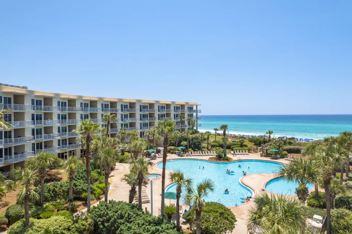 Aerial view of a multi-story condominium building with numerous balconies overlooking a large, freeform swimming pool surrounded by palm trees and lush landscaping, with the turquoise ocean and white sand beach in the background.
