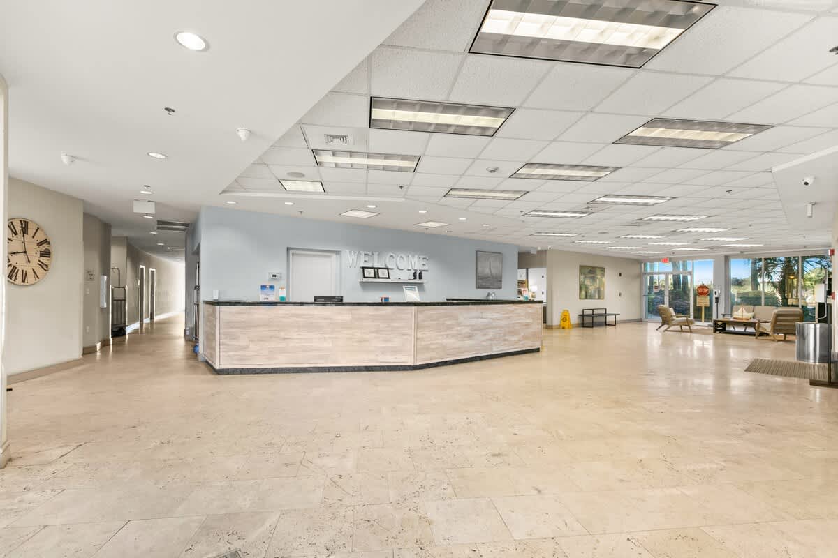 A spacious and brightly lit lobby with light-colored tile flooring, featuring a long reception desk with a "WELCOME" sign. The area includes seating, a large wall clock, and leads to other parts of the building with outdoor views.