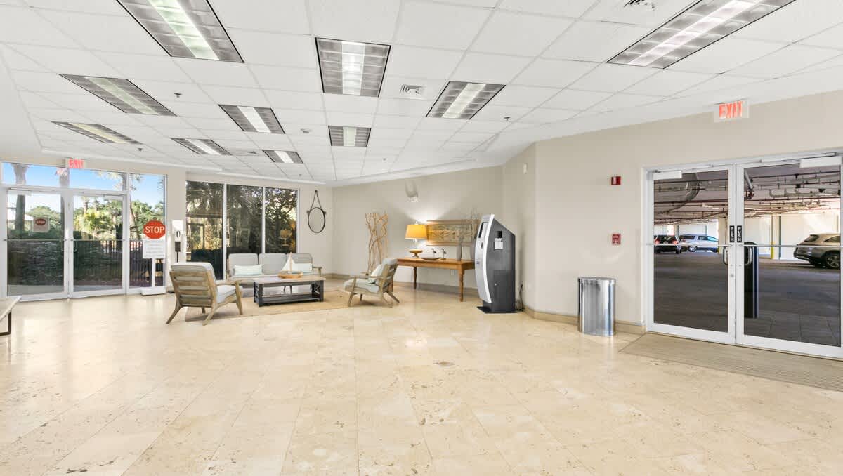A bright foyer with light tiled flooring and a drop ceiling, featuring a cozy seating area with a light-colored sofa, two armchairs, and a coffee table on a rug. Large glass exit doors lead to an outdoor parking area, and a DVD rental machine stands near the wall.